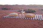 Erg Chigaga Star Camp striped outdoor rug with wooden table, glassware, wine bottle, and two yellow cushions on desert sand