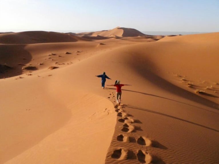 Desert landscape with two people walking along golden sand dunes under clear sky at Erg Chigaga Star Camp