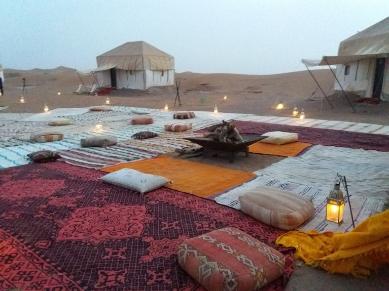 Erg Chigaga Star Camp outdoor seating area with colorful patterned rugs, cushions, lanterns, and tents in desert sand
