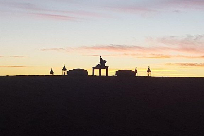 Erg Chigaga Star Camp silhouette with a person sitting on a table at sunset, flanked by lanterns and cushions on sand dunes