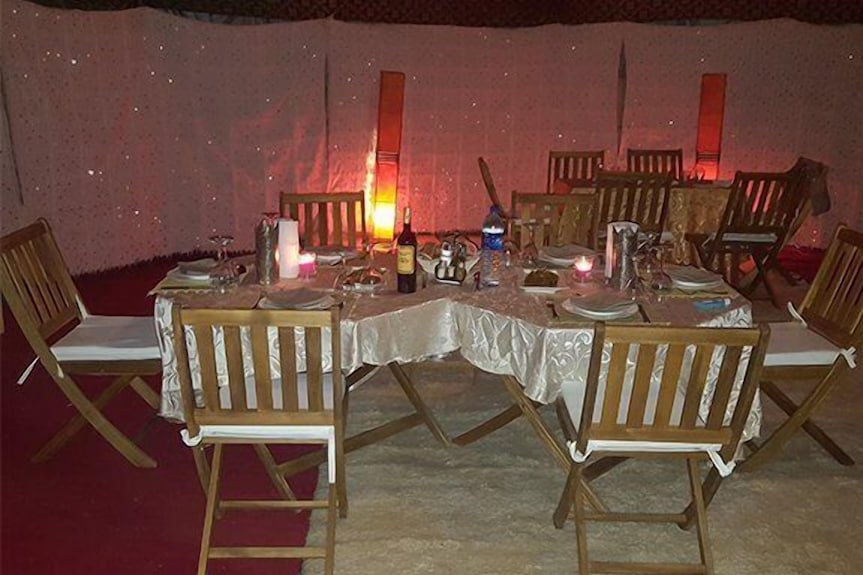 Erg Chigaga Star Camp wooden dining chairs with white cushions around a table covered in a light patterned tablecloth with dim warm lighting