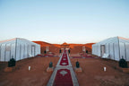 Errachidia Airport desert camp with white canvas tents, red carpet walkway, and lanterns on sandy ground near orange dunes at sunset