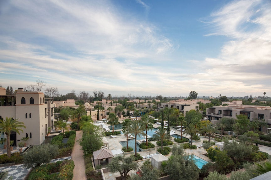 Four Seasons Resort Marrakech luxury hotel complex with beige Moroccan-style buildings, lush green palm trees, landscaped gardens, and swimming pools under a partly cloudy sky