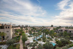Four Seasons Resort Marrakech luxury hotel complex with beige Moroccan-style buildings, lush green palm trees, landscaped gardens, and swimming pools under a partly cloudy sky