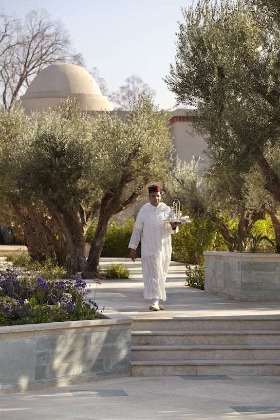 Four Seasons Resort Marrakech with a server in traditional white attire and red hat carrying a tray in a landscaped garden with olive trees and dome architecture