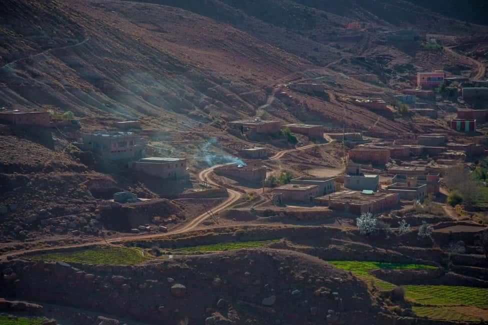 From Marrakech to Ourika Valley tour showing a rugged Atlas Mountain village with earthen homes, winding roads, and green fields
