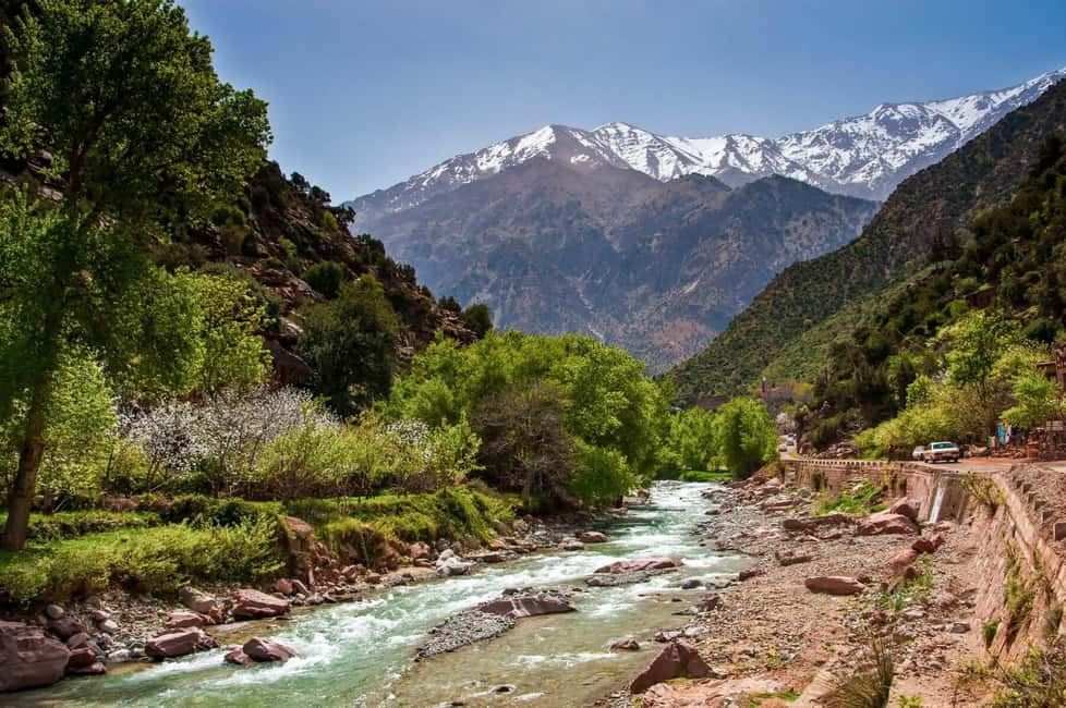 From Marrakech to Ourika Valley tour featuring a turquoise river, lush green valley, rocky banks, and snow-capped Atlas Mountains