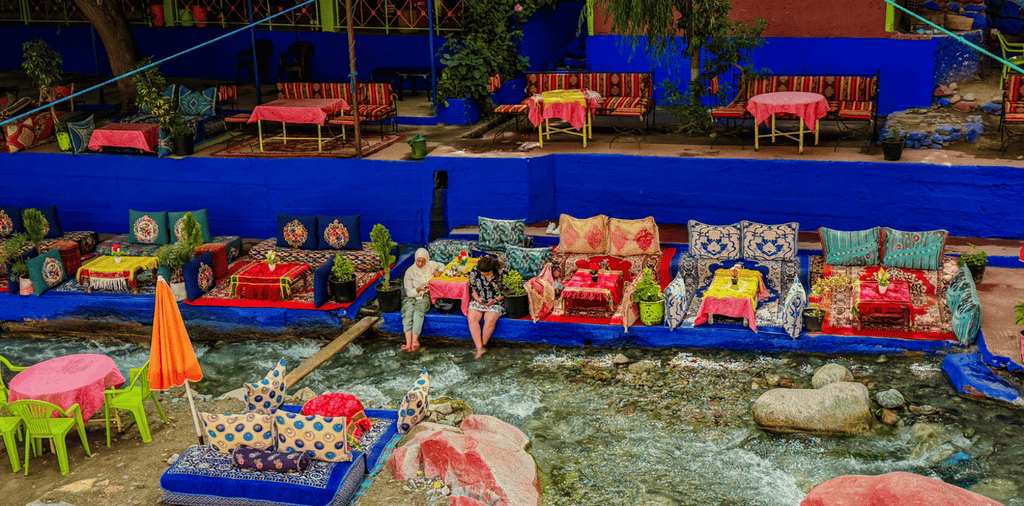From Marrakech to Ourika Valley tour showing colourful riverside cushions, tables, umbrellas and flowing mountain stream