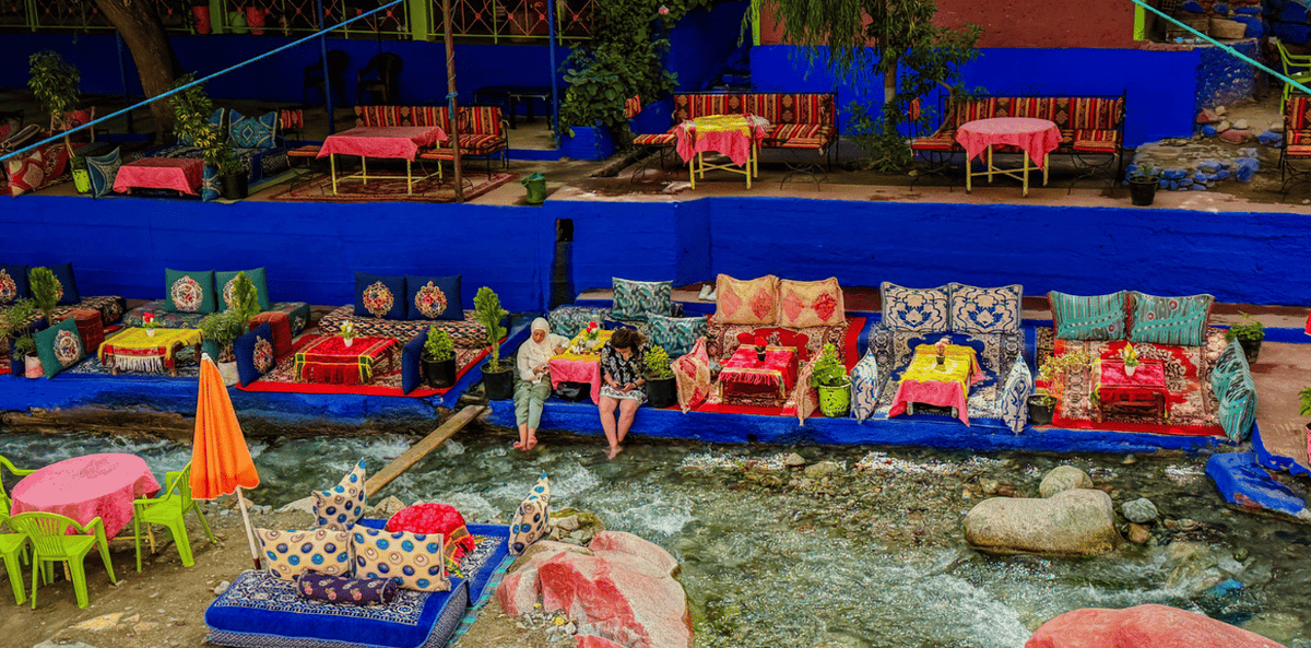 From Marrakech to Ourika Valley tour showing colourful riverside cushions, tables, umbrellas and flowing mountain stream