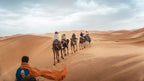 From Marrakech: To Sahara Tour with camel riders crossing orange desert dunes, led by a blue-robed guide