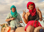 From Marrakech: To Sahara Tour featuring two travellers in colorful desert clothing riding a patterned camel saddle