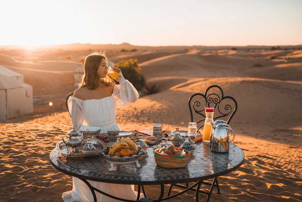 From Marrakech: To Sahara Tour featuring a woman enjoying Moroccan tea at a desert table with pastries
