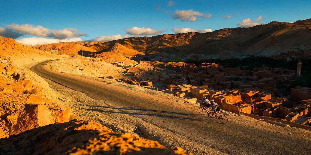 From Marrakech: To Sahara Tour showing a winding mountain road, ochre village, and two cyclists in Morocco