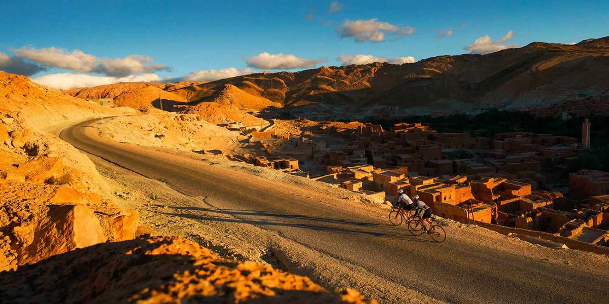 From Marrakech: To Sahara Tour showing a winding mountain road, ochre village, and two cyclists in Morocco