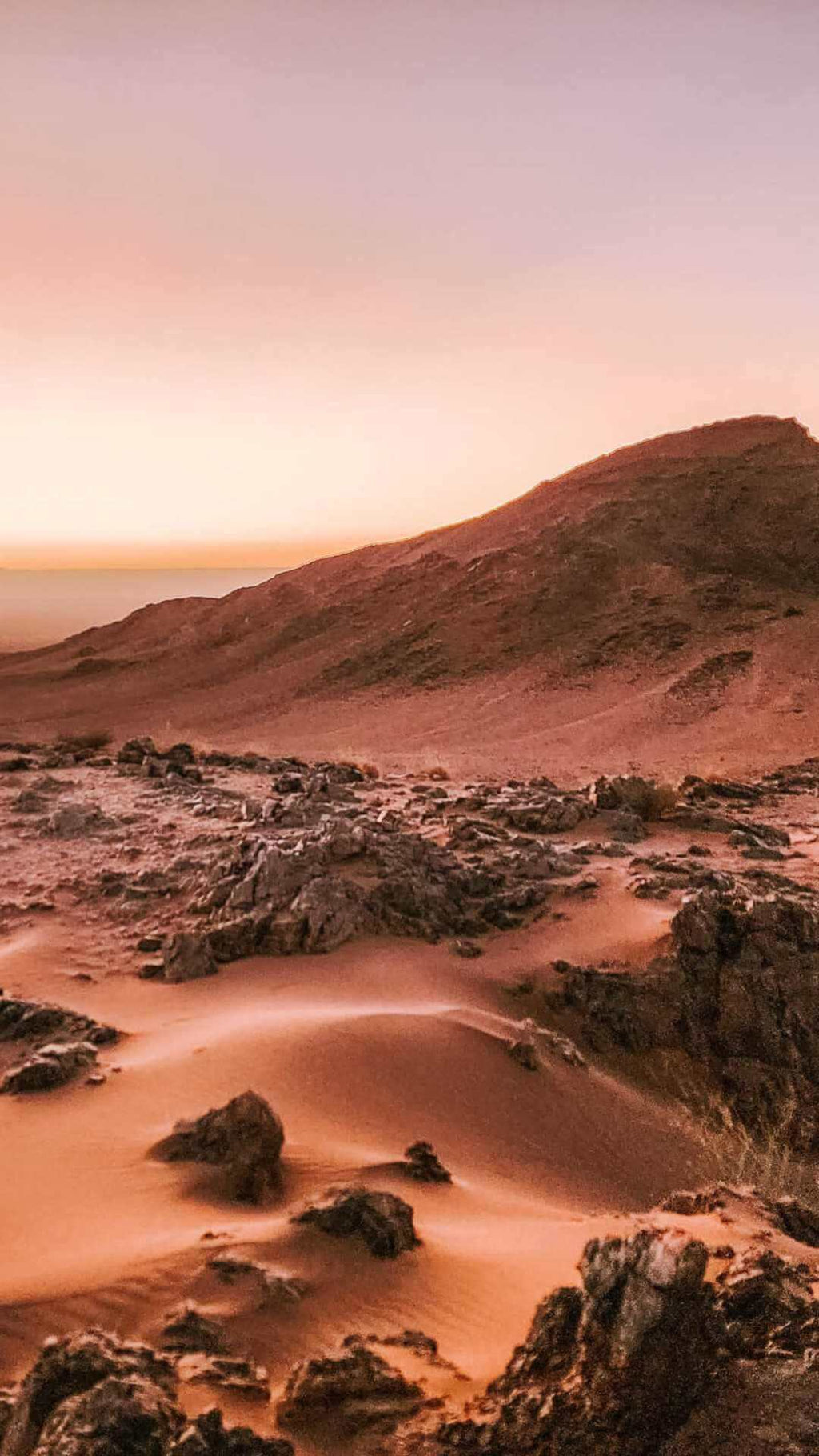 From Marrakech: To Sahara Tour showing red desert dunes with dark rocky formations beneath a pink sky