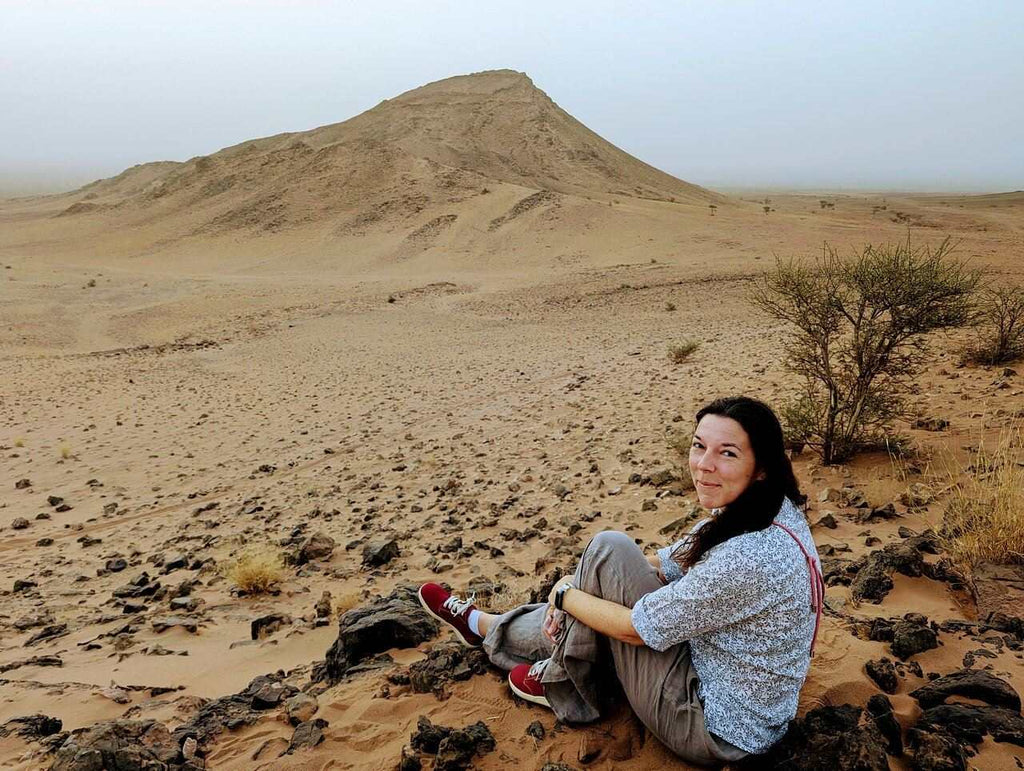 From Marrakech: To Sahara Tour with woman seated on rocky desert terrain beneath a sandy mountain