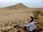 From Marrakech: To Sahara Tour with woman seated on rocky desert terrain beneath a sandy mountain