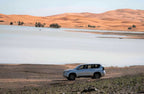 From Marrakech: To Sahara Tour featuring a silver SUV beside a lake, with orange desert dunes and sparse palms