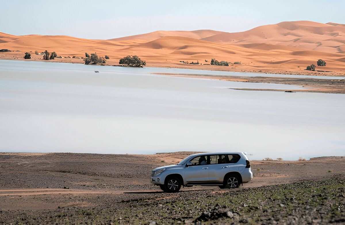 From Marrakech: To Sahara Tour featuring a silver SUV beside a lake, with orange desert dunes and sparse palms