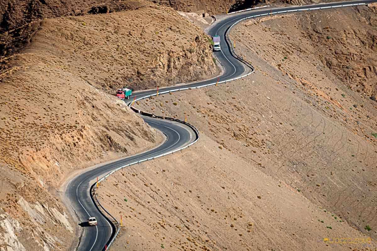 From Marrakech: To Sahara Tour through a winding mountain road with guardrails and vehicles across arid rocky terrain