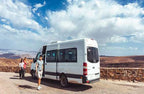 White passenger van parked near stone wall with three casually dressed travelers walking beside it under partly cloudy blue sky, representing Group Tours from Marrakech.
