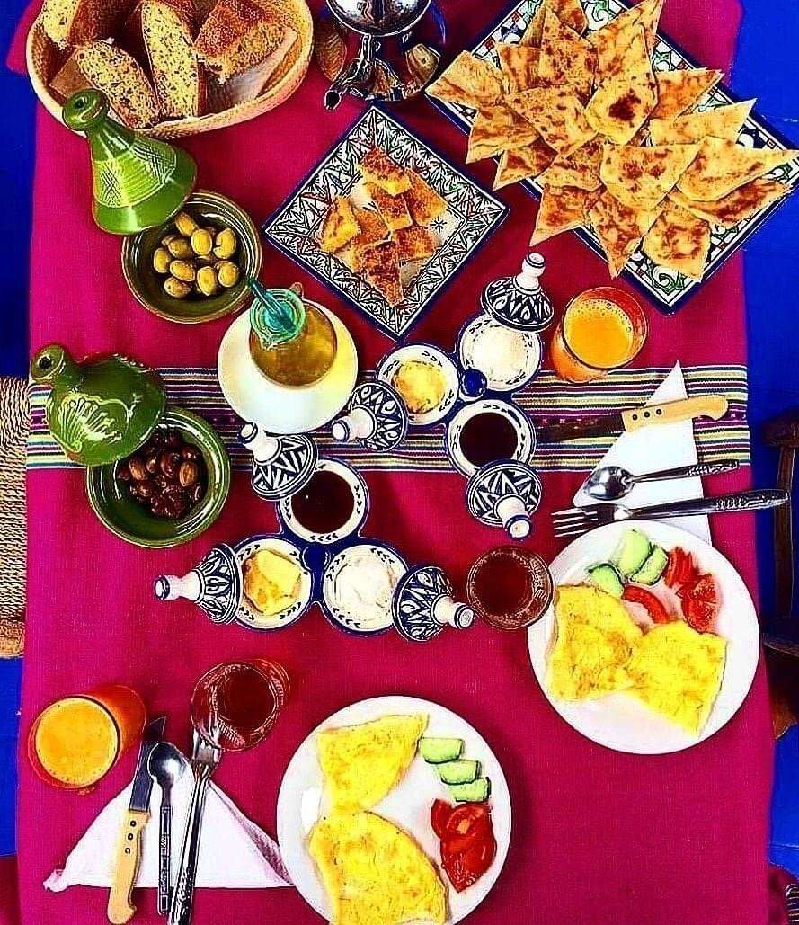Traditional Moroccan breakfast spread with omelettes, sliced cucumbers and tomatoes, flatbread, olives, orange juice, and decorative ceramic dishes on a vibrant pink tablecloth, showcasing Group Tours from Marrakech hospitality.