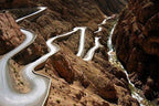 Winding paved mountain road with sharp hairpin turns through rocky brown cliffs and a narrow river below for Group Tours from Marrakech.