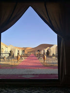 Desert camp entrance with beige tents, red carpets, metal chairs, and large clay pots against clear blue sky and sand dunes for Group Tours from Marrakech.