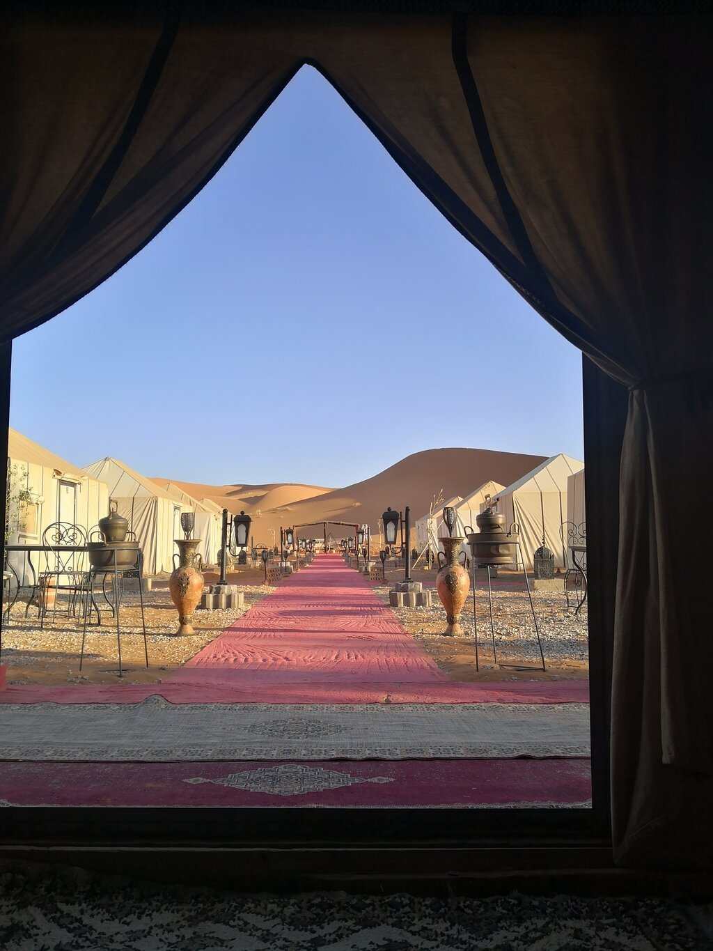 Desert camp entrance with beige tents, red carpets, metal chairs, and large clay pots against clear blue sky and sand dunes for Group Tours from Marrakech.