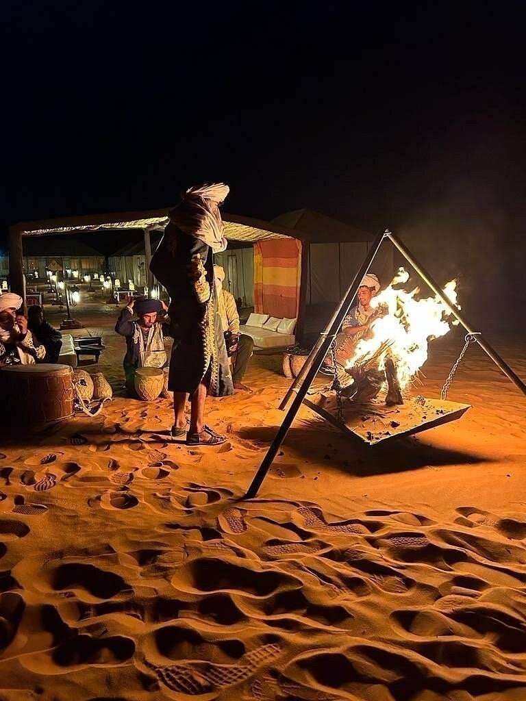 Desert camp scene at night with traditional Moroccan musicians and dancers around an open fire on sandy ground, warm tones. Group Tours from Marrakech.