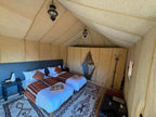 Tent interior with twin beds dressed in white sheets and patterned rust-orange blankets, Moroccan rugs, and a small bedside table and coat rack, cozy and rustic for Group Tours from Marrakech.