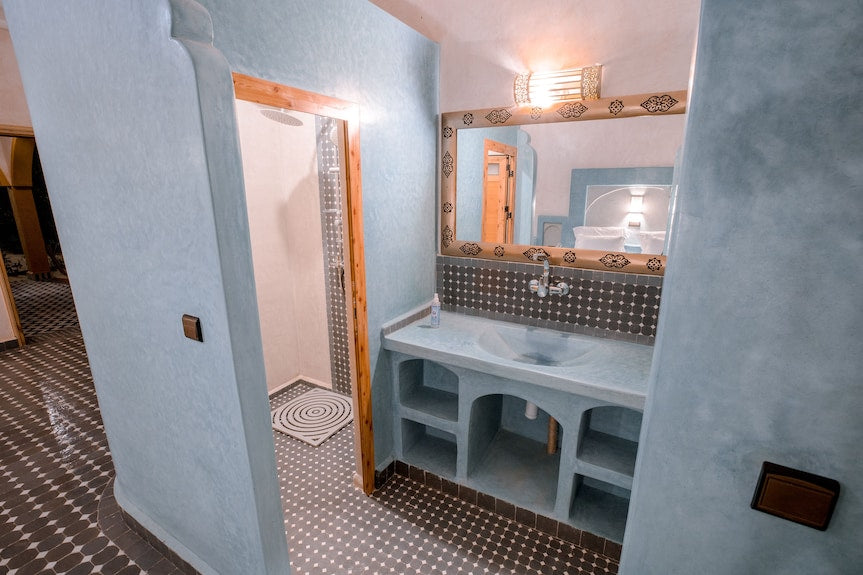 Bathroom with blue plastered walls, patterned brown and white floor tiles, wooden door frame, and a mirror above the sink at Hassilabiad Appart Hotel