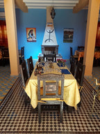 Dining area at Hassilabiad Appart Hotel with wooden chairs, yellow tablecloth, tiled floor, and a decorative fireplace on blue wall