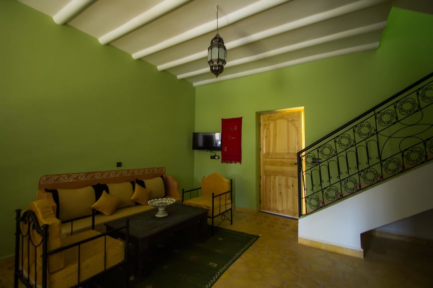 Hassilabiad Appart Hotel living room with green walls, wooden ceiling beams, wrought iron staircase, and mustard cushioned seating