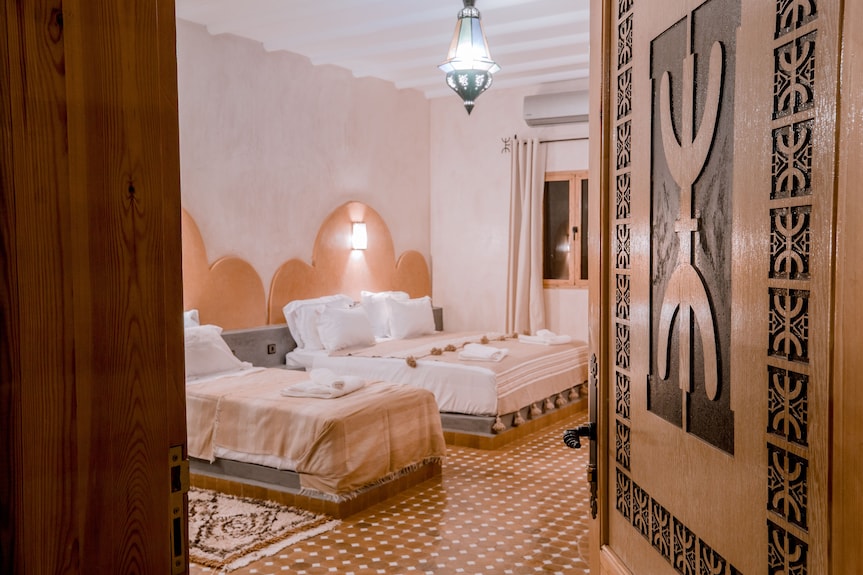 Hassilabiad Appart Hotel room with two beige beds, white linens, Moroccan style decor, patterned tile floor, and wooden door with carved design