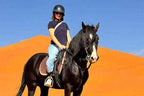 Woman wearing a black helmet and navy shirt riding a black horse with white markings on its face in front of orange sand dunes, illustrating Horse Riding in Merzouga.