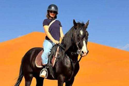 Woman wearing a black helmet and navy shirt riding a black horse with white markings on its face in front of orange sand dunes, illustrating Horse Riding in Merzouga.
