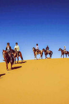 Group of riders on brown horses crossing golden sand dunes under clear blue sky during Horse Riding in Merzouga desert adventure