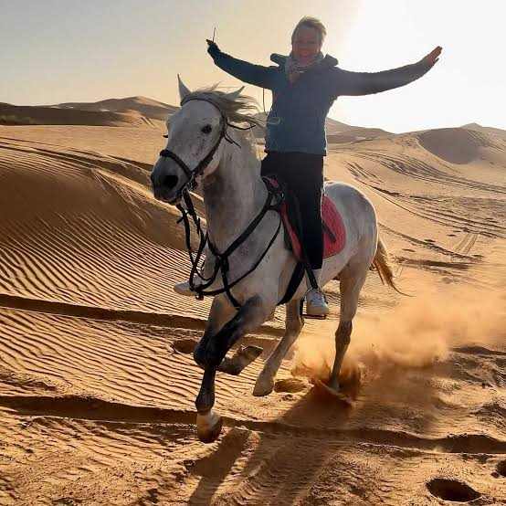 White horse with pink saddle pad galloping on sandy desert dunes with a rider raising arms, showcasing Horse Riding in Merzouga.