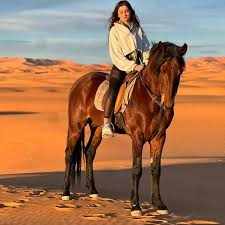 Horse Riding in Merzouga featuring a rider wearing a white jacket on a brown horse against the orange Sahara dunes backdrop