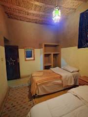 Hostel Dar Lgamra bedroom with beige walls, two single beds, wooden ceiling, patterned rug, and colorful hanging light