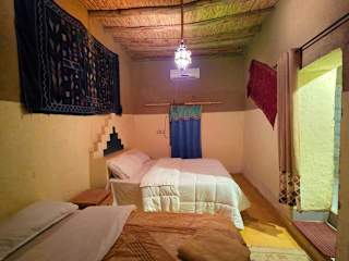 Hostel Dar Lgamra bedroom with two single beds, white bedding, Moroccan rugs on walls, beige walls, and a hanging light fixture