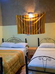 Hostel Dar Lgamra bedroom with two metal-framed twin beds, yellow and white bedding, and a yellow striped textile wall hanging