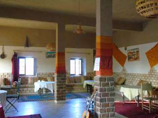 Hostel Dar Lgamra interior with colorful rugs on columns and windows, wooden furniture, and chairs creating a cozy atmosphere