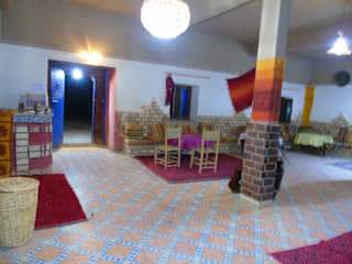 Hostel Dar Lgamra interior with tiled floor, wooden chairs and tables, colorful rugs, and decorative brick pillars