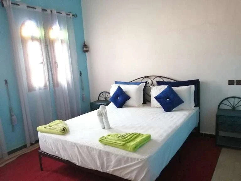 Hotel auberge Sahara bedroom with white bed linens, blue and white pillows, green towels, blue walls, and arched windows with white curtains