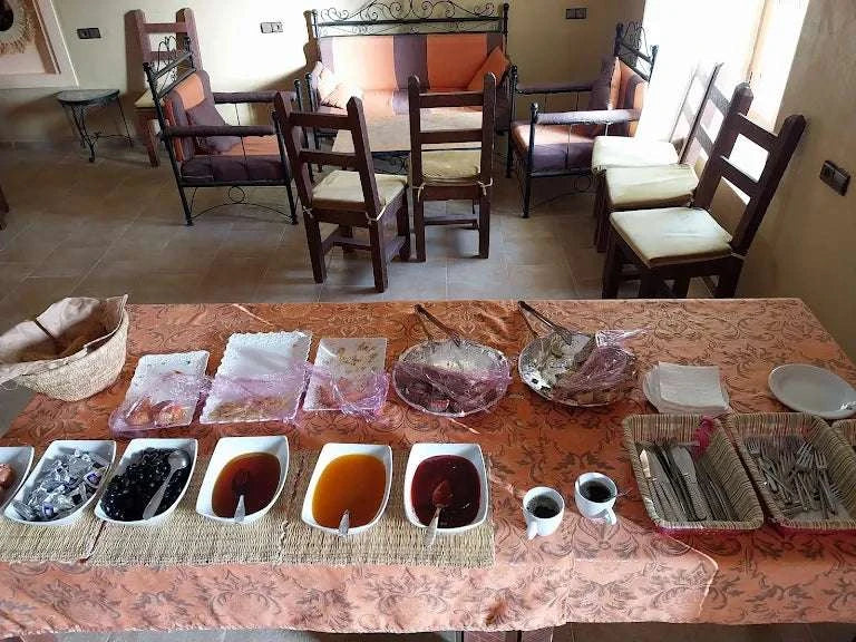 Hotel auberge Sahara breakfast table with jars of jam, honey, olives and cups in a room with wooden chairs and orange cushions