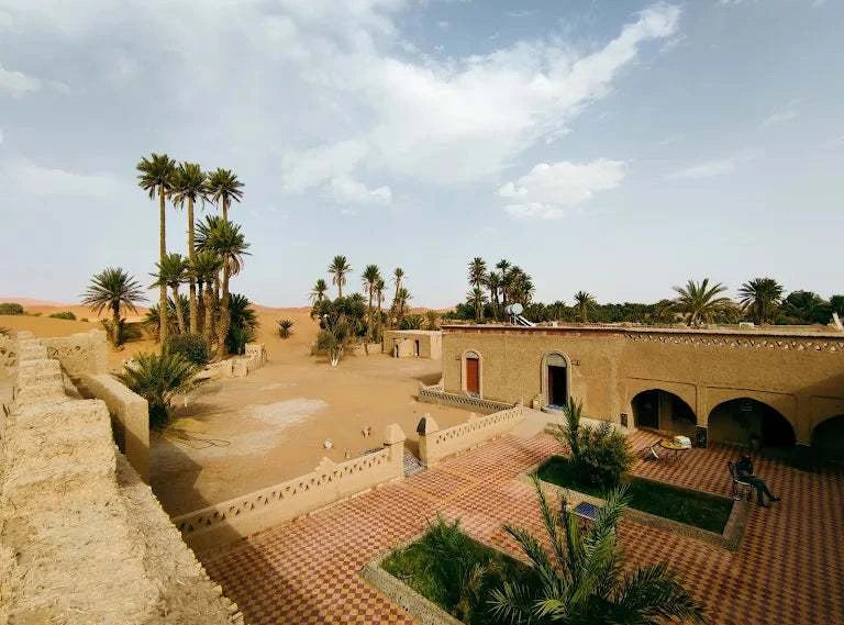 Hotel auberge Sahara exterior view in sandy desert with beige stucco walls, tiled courtyard, and palm trees under cloudy sky