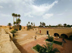 Hotel auberge Sahara exterior view in sandy desert with beige stucco walls, tiled courtyard, and palm trees under cloudy sky
