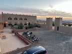 Hotel Camping Les Roses featuring traditional beige adobe buildings with decorative crenellations and a motorcycle parking area under a colorful sunset sky